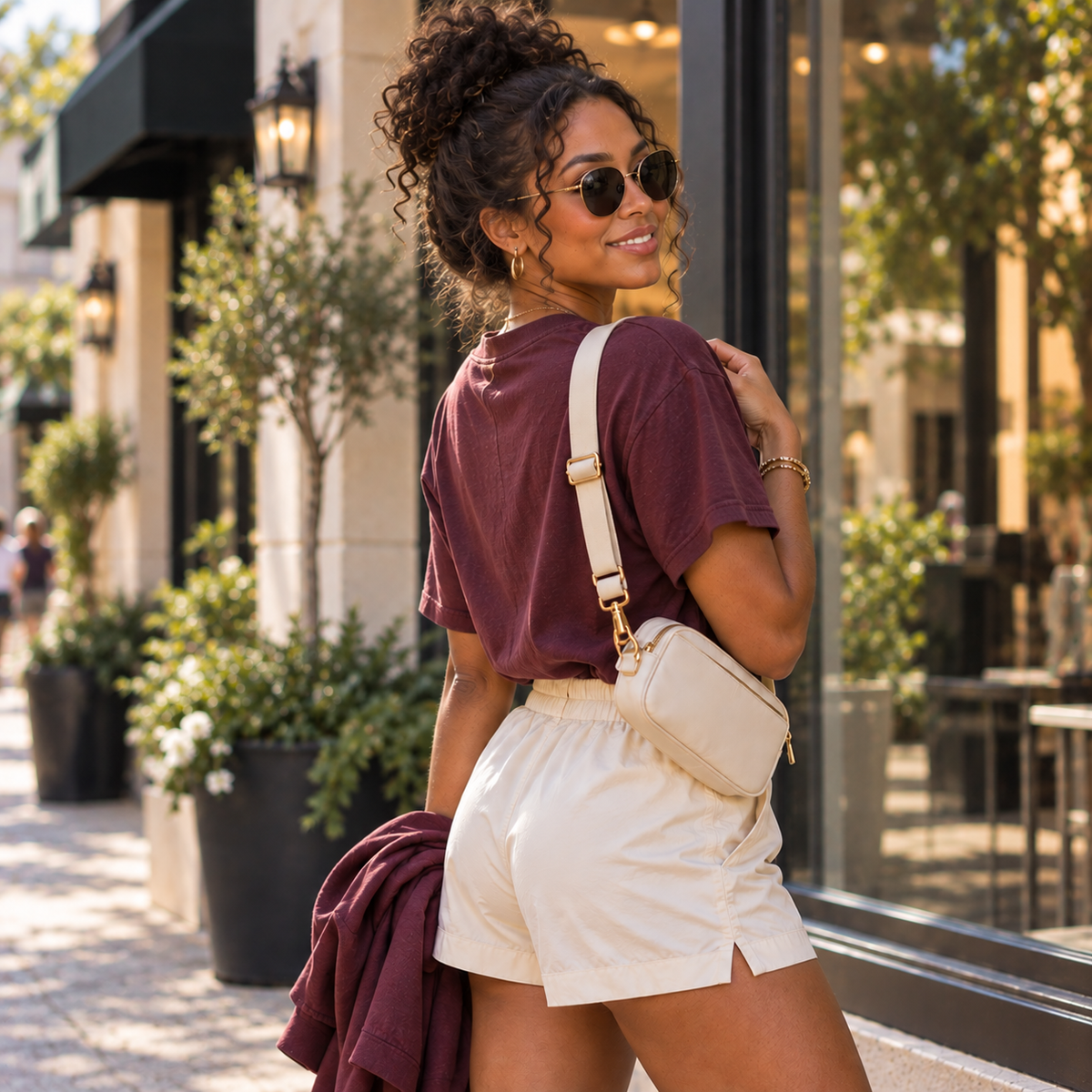 A woman wearing a maroon t-shirt, cream shorts, and a cream crossbody bag stands on a sunny city sidewalk in front of storefront windows.