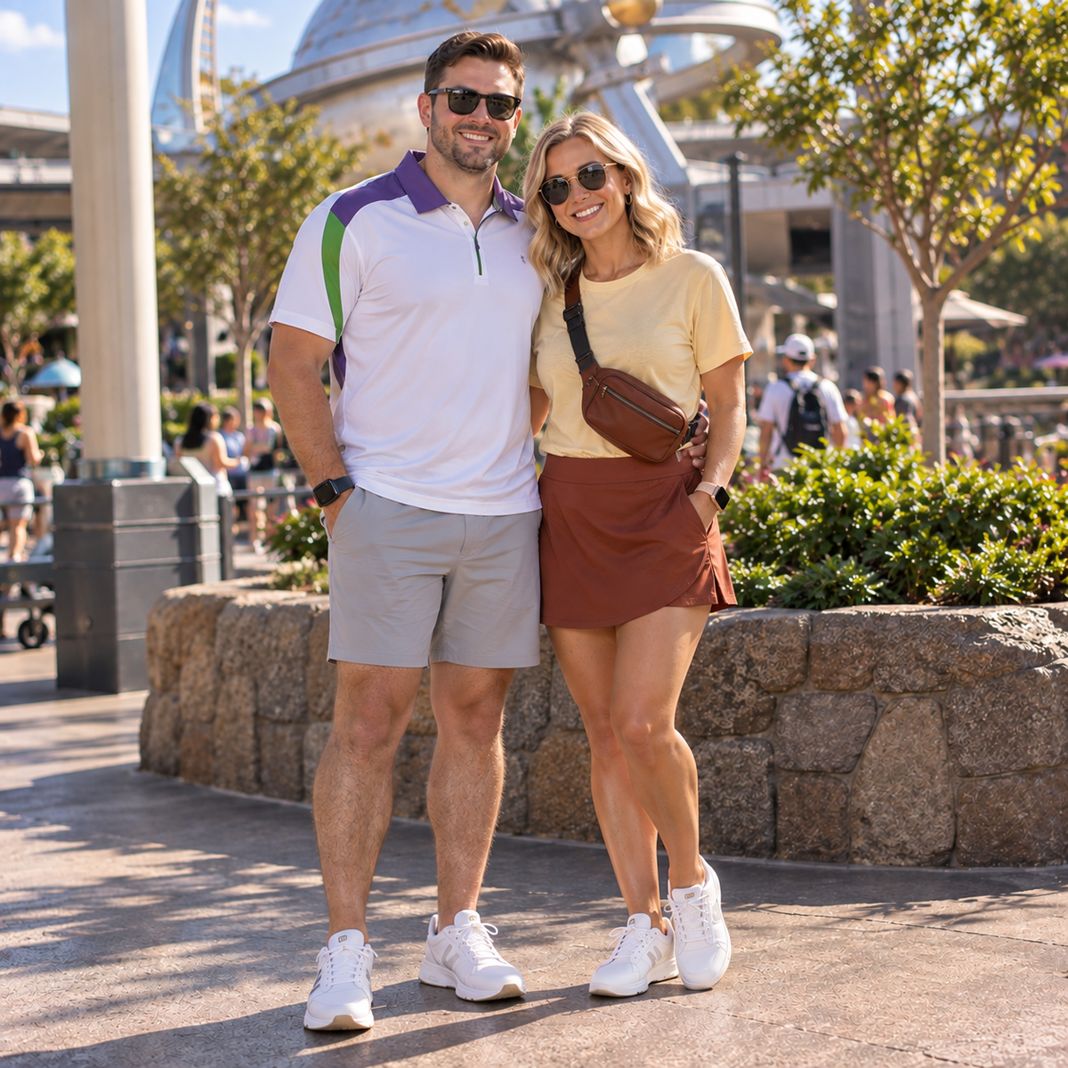 A man and a woman standing together outdoors in a park-like setting with a large domed structure in the background. The man wears a white polo shirt with purple and green accents, gray shorts, and white sneakers.