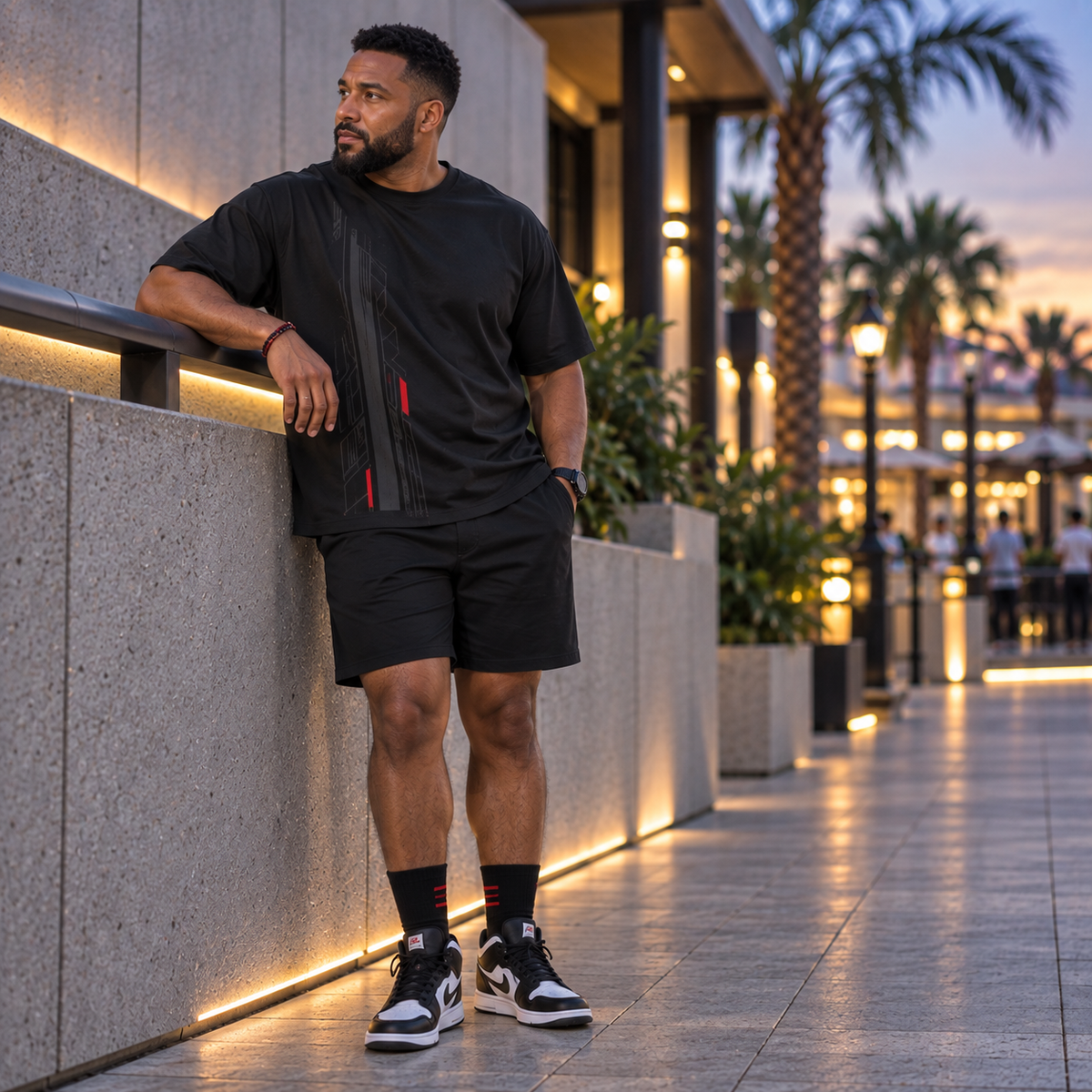 A man wearing a black short-sleeve zip-front t-shirt and matching black shorts stands leaning against a modern stone wall in an outdoor walkway at dusk.